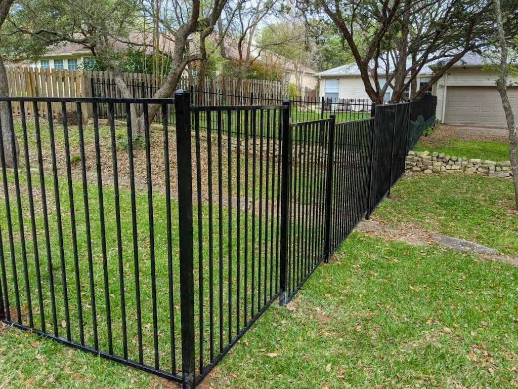 A black metal fence runs through a green lawn, bordered by trees. A wooden fence and garage are in the background, conveying a suburban feel.