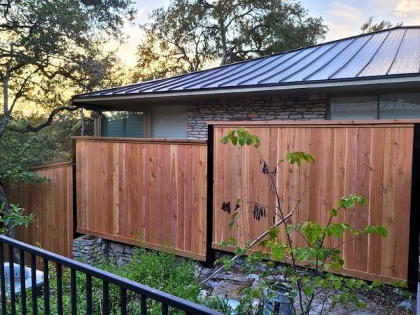 wooden privacy fence with a natural finish surrounds a stone house amid lush greenery. A serene sunrise or sunset lights up the sky, creating a calm ambiance.