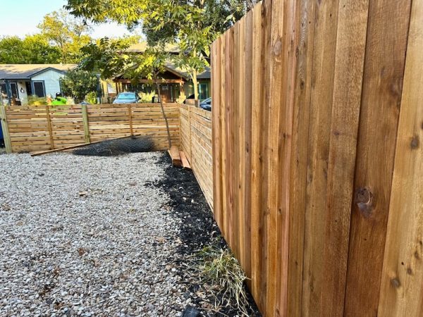 wooden privacy fence and gravel area in a backyard. A chain-link roll and wood planks lie next to the fence. Houses and trees are visible in the background.