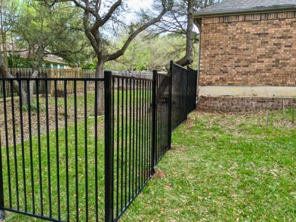 black metal fence bending sharply around a brick house, set against a backdrop of trees and lush grass, conveying a serene suburban atmosphere.