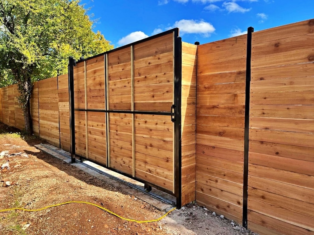 Wooden privacy fence with a sliding gate, framed in black metal, against a clear blue sky and lush green trees. A yellow hose lies on the ground.