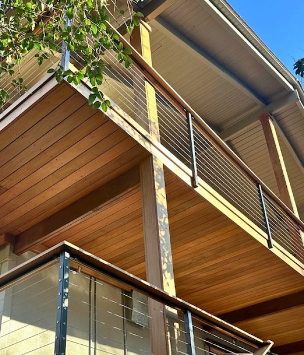 Two-story wooden house with modern, open balconies featuring cable railings. Overhanging eaves and surrounding green foliage convey a serene setting.