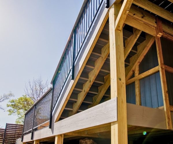 Low angle view of a wooden deck with black metal railing under a clear blue sky. Trees and a wooden privacy fence surround the structure.