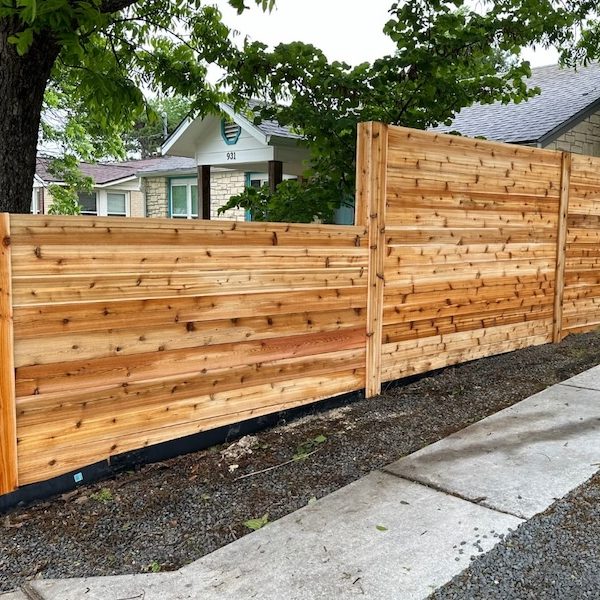 new wooden fence with horizontal planks lines a sidewalk, bordered by a lush green tree and a suburban house. The scene feels peaceful and tidy.