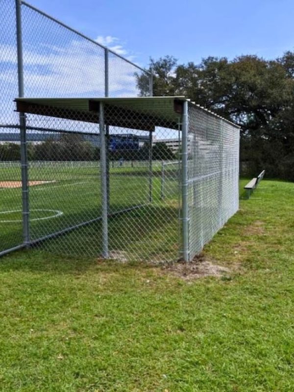 Chain-link fence structure shelters a baseball field with bases marked on green grass. Bright sky and trees form a serene backdrop.