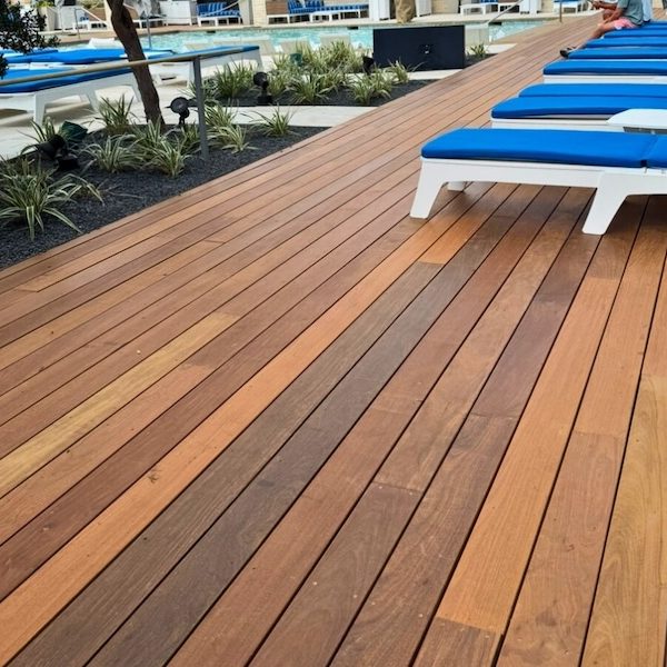 Wooden deck by a pool lined with blue-cushioned white lounge chairs. Fresh greenery borders the area, adding a tranquil, relaxing atmosphere.