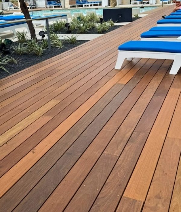 Wooden deck by a pool lined with blue-cushioned white lounge chairs. Fresh greenery borders the area, adding a tranquil, relaxing atmosphere.