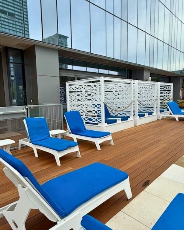 Poolside area with blue cushioned lounge chairs on a wooden deck beside a modern building. The scene is calm and inviting, suggesting relaxation.