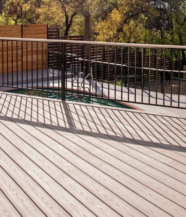 Sunlit wooden deck with black metal railing casting shadows. A pool is partially visible, surrounded by tall wooden privacy fencing and lush trees.