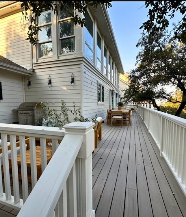 Expansive wooden deck attached to a light gray house, featuring outdoor furniture, a grill, and a TV. Surrounded by lush trees, creating a serene setting.