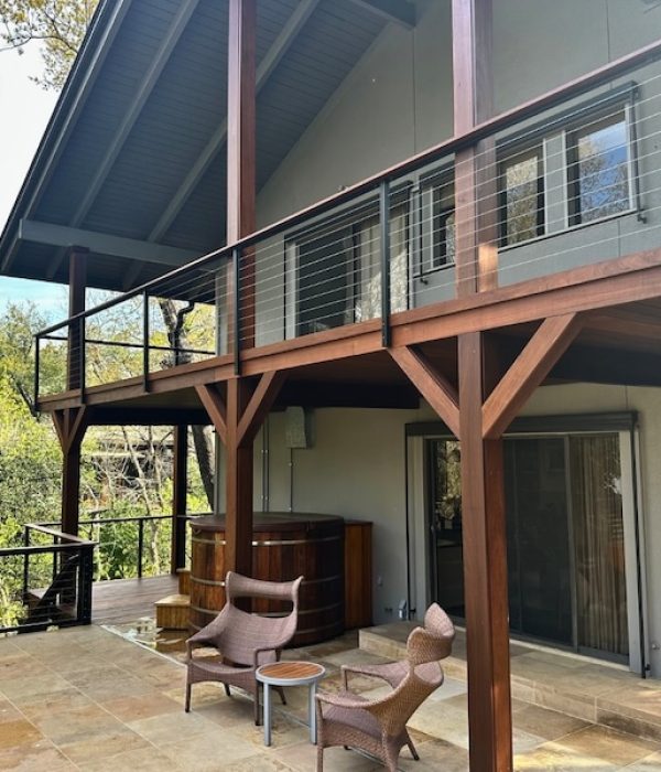 Two-story house with a wooden deck, wicker chairs, and a hot tub surrounded by lush trees. The scene is serene and inviting under a clear sky.