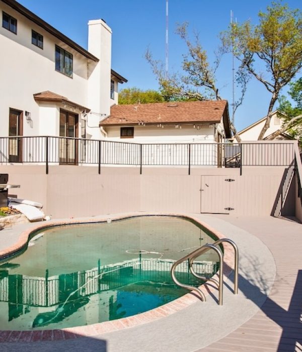 Backyard view with a clean pool surrounded by a concrete deck and black metal railing. A barbecue grill and a white house with stairs in the background. Sunny day.