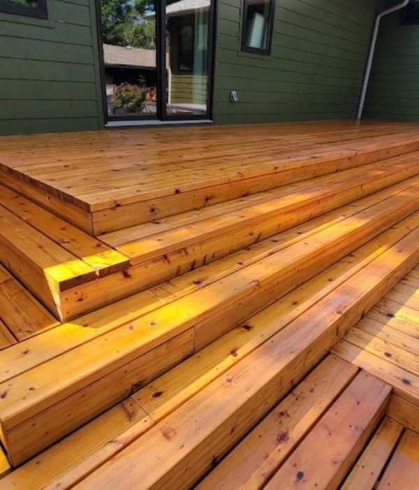 Brightly lit wooden deck with wide, tiered steps leading up to a green house. The warm wood tones evoke a relaxed, inviting atmosphere.