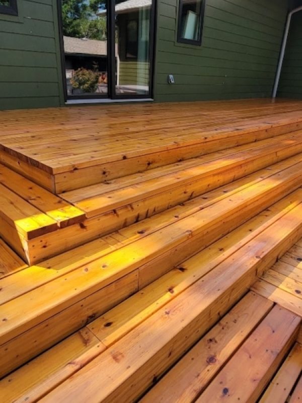 Brightly lit wooden deck with wide, tiered steps leading up to a green house. The warm wood tones evoke a relaxed, inviting atmosphere.