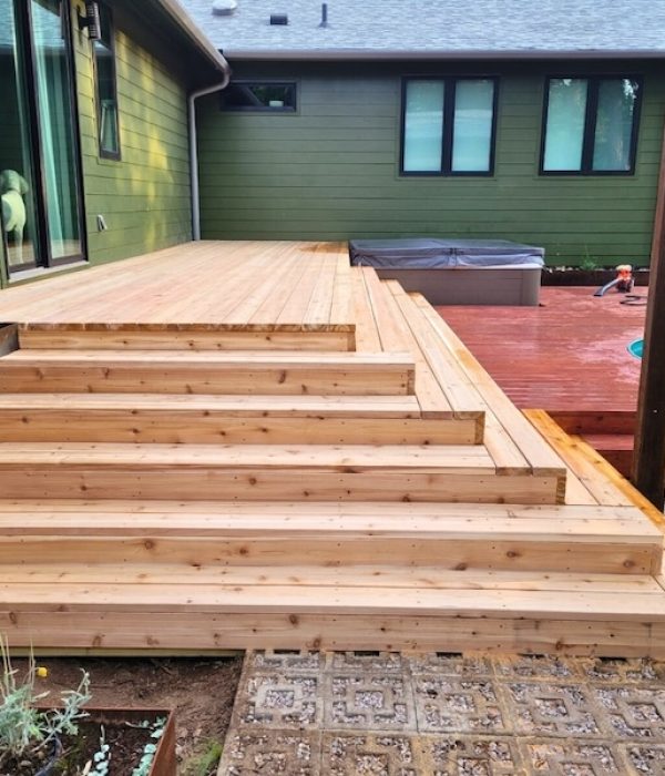 A wooden deck with wide steps leads to a patio area next to a house with green siding. The patio features a hot tub and a pool. A person stands nearby.