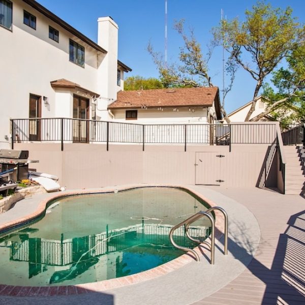 Backyard view with a clean pool surrounded by a concrete deck and black metal railing. A barbecue grill and a white house with stairs in the background. Sunny day.