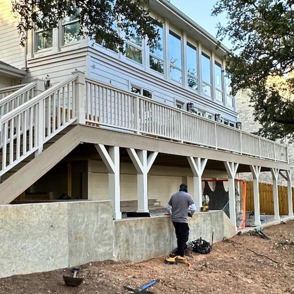 Two-story house with a large deck featuring white railings and stairs. A person works under the deck; the yard has tools and soil ready for landscaping.