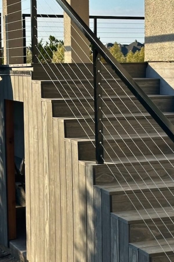 A worker stands on a ladder, working on a wooden structure next to a staircase with a sleek metal railing. The scene is bathed in warm sunlight.