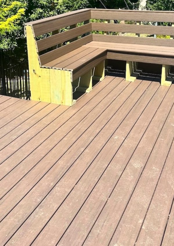 A wooden deck with built-in corner seating and black metal railings. It overlooks greenery, including bright yellow shrubs, on a sunny day.
