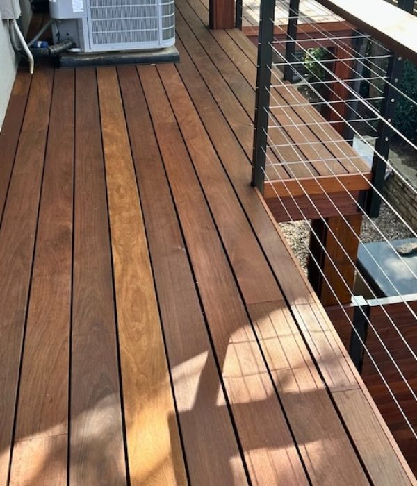 Wooden deck with smooth, brown planks. A metal cable railing runs along the edge, offering a view of a garden below with bushes and pebbles.