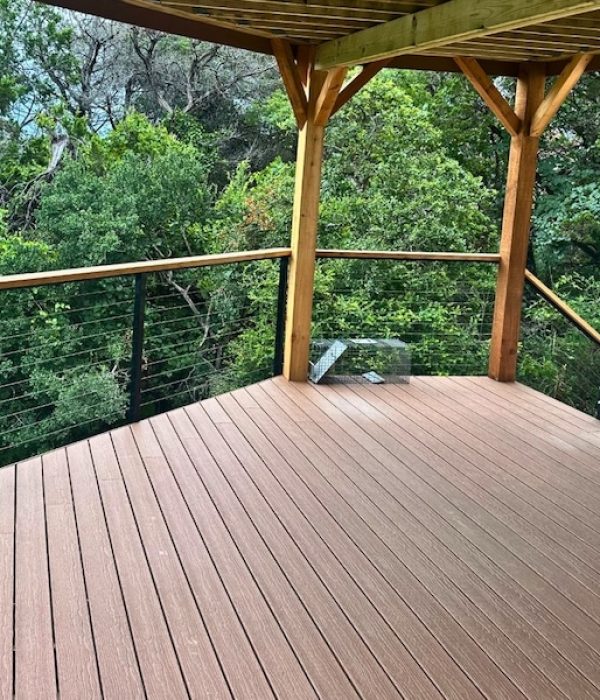 Wooden deck with railing overlooks lush green trees. The scene conveys tranquility and connection with nature, under a partially covered area.