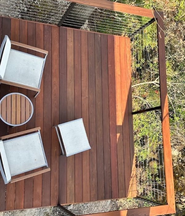 Aerial view of a wooden deck with two white cushioned chairs, a small round table, and a footrest. Surrounded by a metal railing and lush green trees.