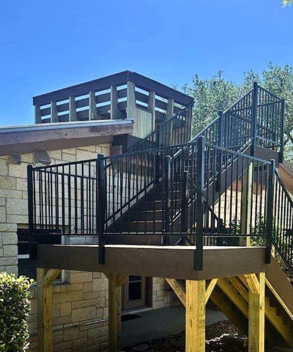 Outdoor staircase with black railings leads to a modern rooftop deck on a stone building. Lush greenery and a clear blue sky set a serene tone.