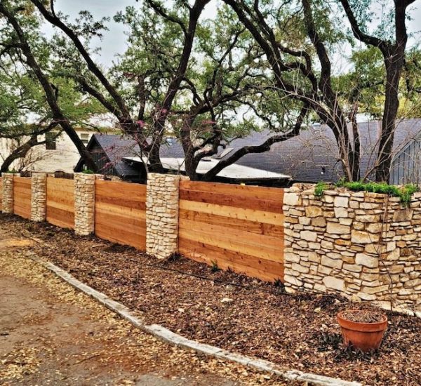 rustic wooden fence with stone pillars borders a yard with large, leafy trees in the background. The scene exudes a calm and natural atmosphere.
