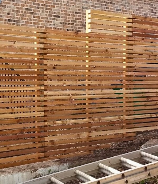 Wooden slatted privacy fence in front of a brick wall, with an aluminum ladder on the ground. The scene appears outdoor and work related.
