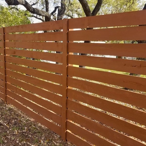 wooden privacy fence with horizontal slats, stained in a warm brown hue, is set against a backdrop of lush greenery and tall trees, evoking a serene and natural atmosphere.