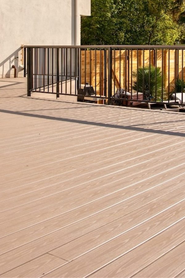 Spacious wooden deck with a sleek metal railing, next to a modern house. Sunlit with few outdoor plants and a wooden fence in the background.