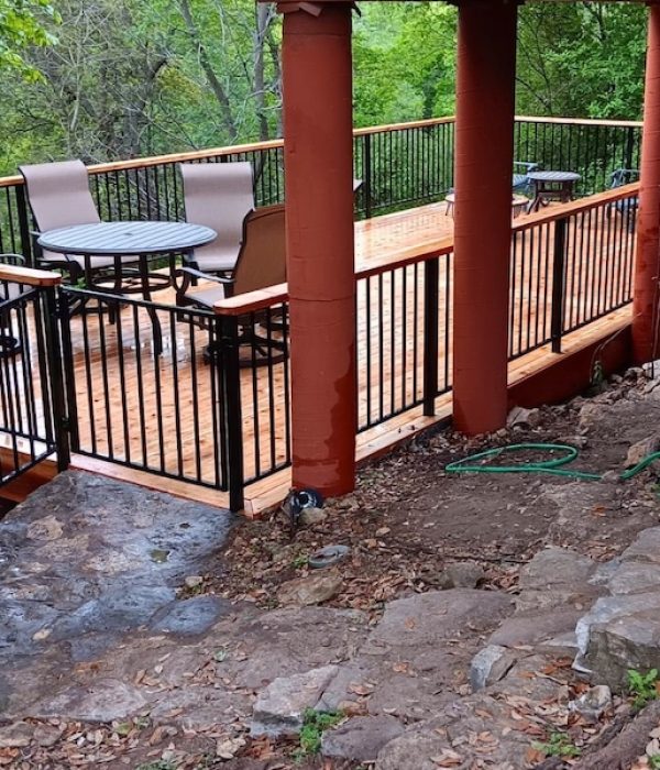 Wooden deck with black railings, set above a rocky slope, features a round table with chairs. Lush green trees provide a serene backdrop.