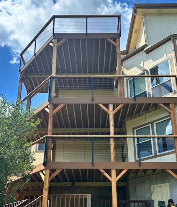 Three-tier wooden deck attached to a beige house under a blue sky with clouds. The structure features cable railings and is surrounded by lush greenery.