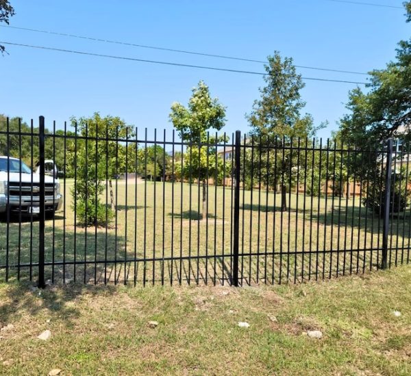 Black metal fence with a closed gate in a grassy yard, flanked by trees under a clear blue sky. A white truck is parked on the left side.